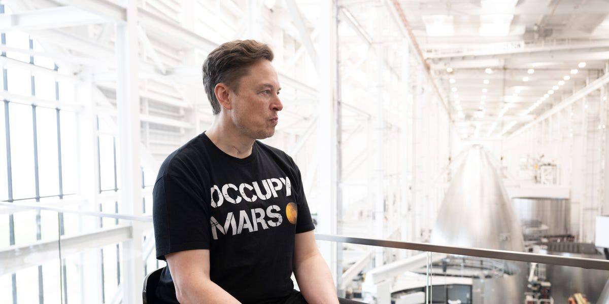 Elon Musk in an "Occupy Mars" t-shirt at the SpaceX factory, with a Starship hull behind him.